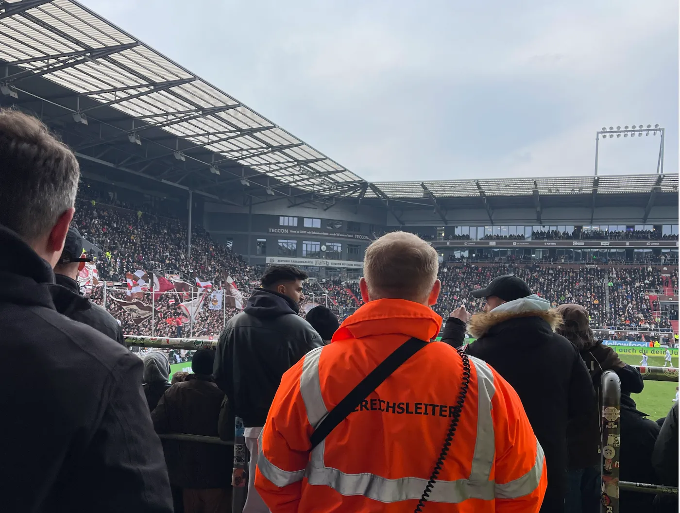 Ordnungsdienst im Fußballstadion Ordnungsdienst in orangefarbener Warnjacke auf der Tribüne eines vollen Fußballstadions