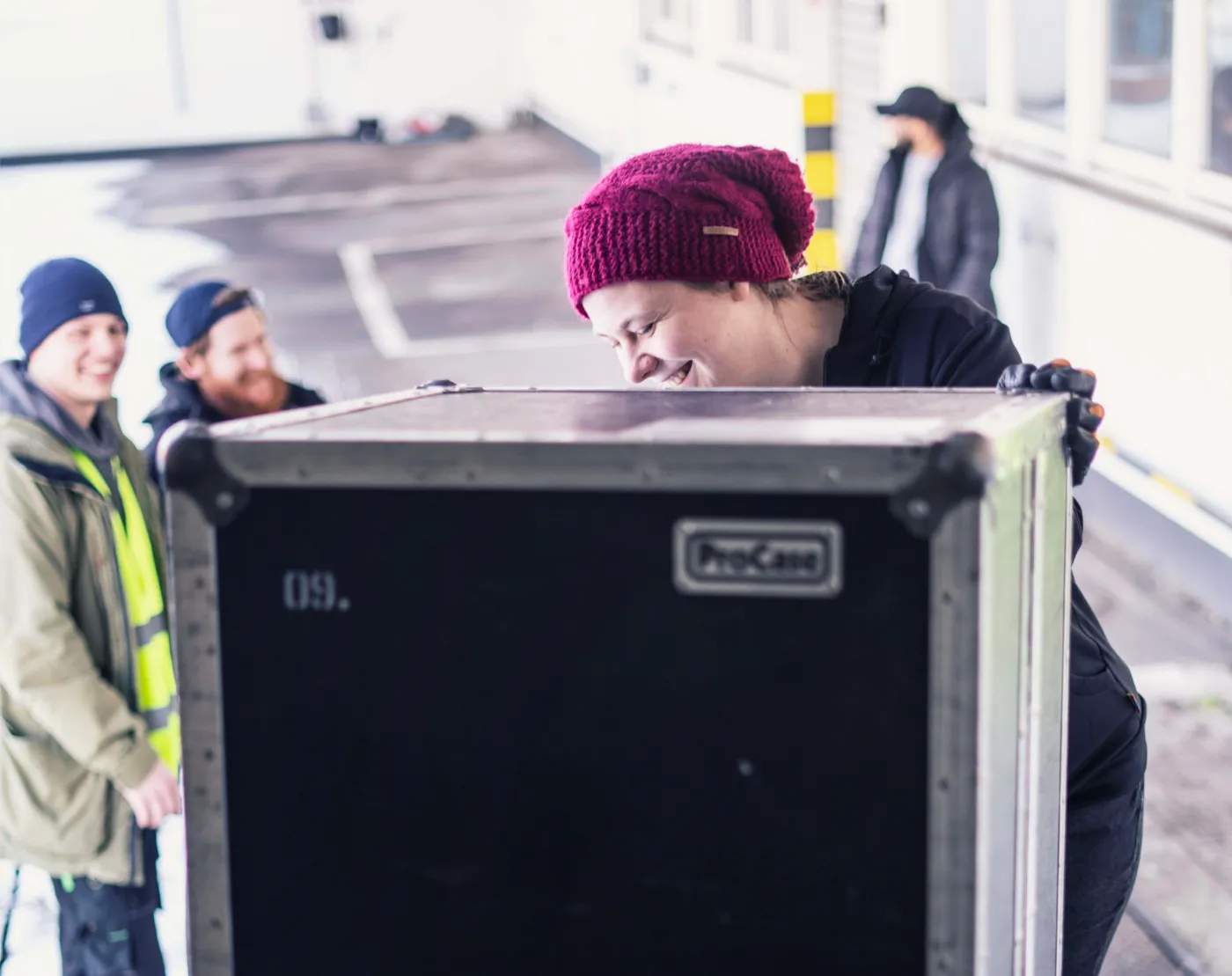 Crew mit Flightcase Crew-Mitglied mit roter Mütze schiebt lachend ein Flightcase, daneben zwei Kollegen