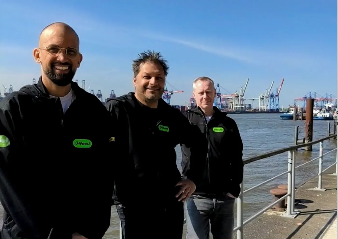 U-Need am Hafen Drei Männer in U-Need-Jacken am Hafen, im Hintergrund Containerkräne