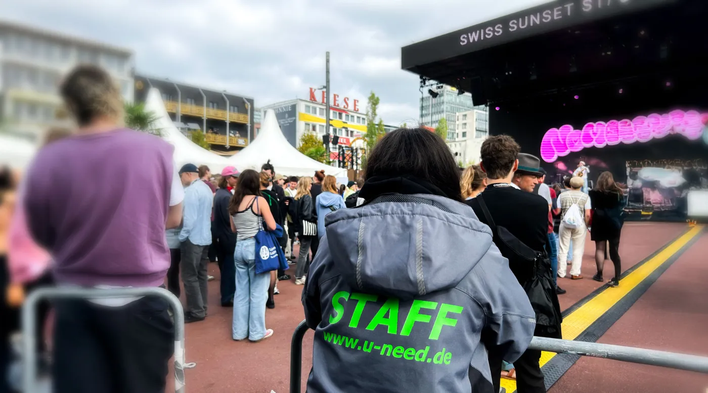 U-Need Staff von hinten neben einer Open-Air-Bühne, davor Besucher